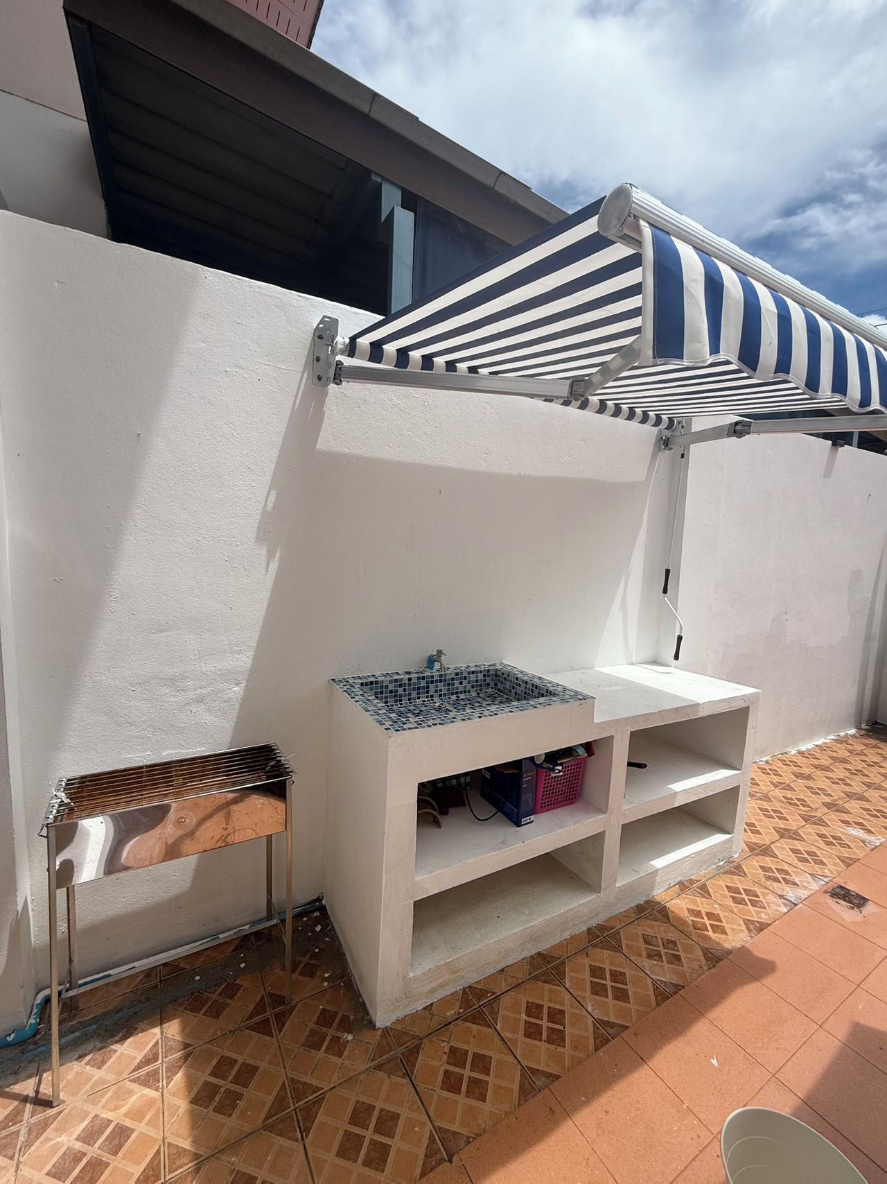 A patio area with a white wall, a striped awning, a built-in sink, and a small bench.