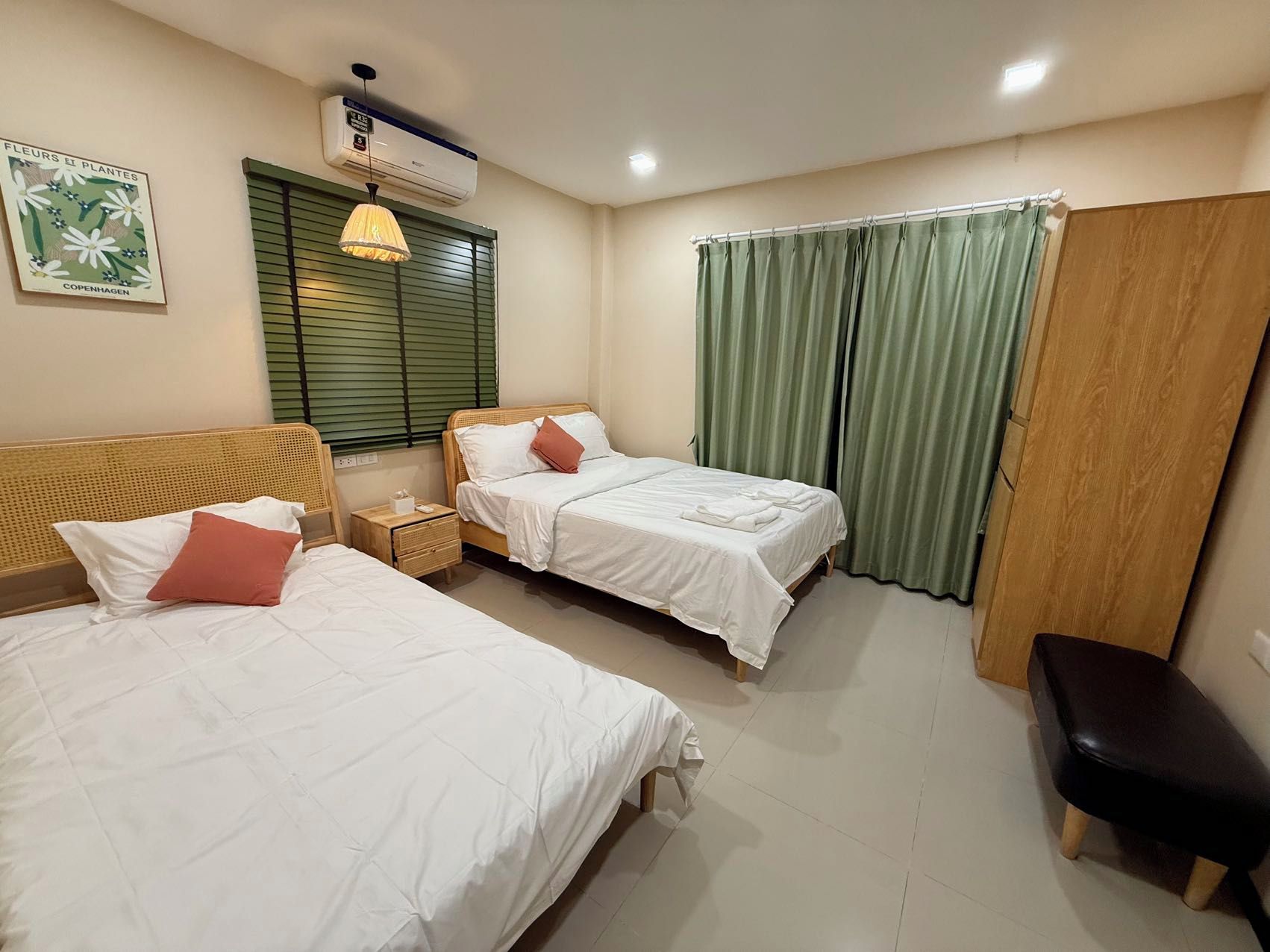 A room with two beds, a nightstand, a bench, a window with blinds, a curtain, and an air conditioning unit.