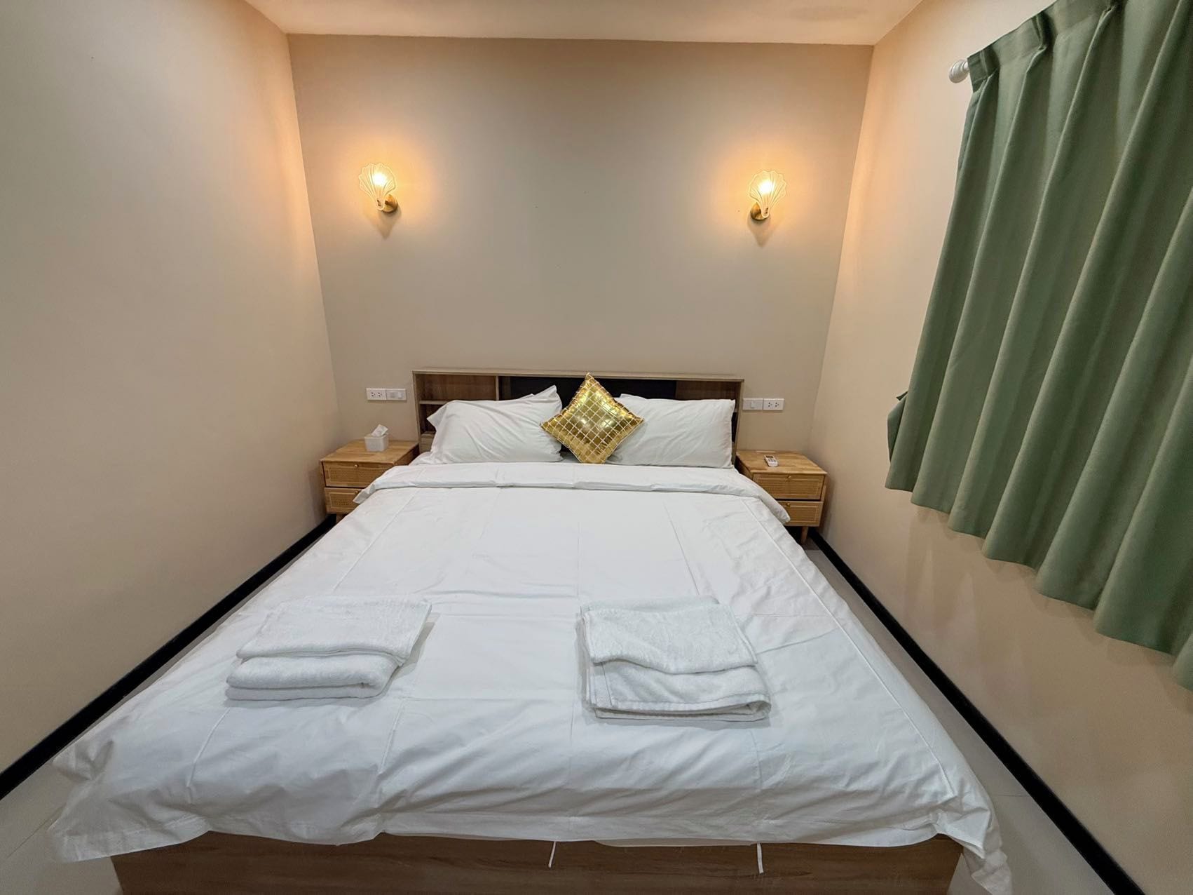 Un lit bien fait, avec du linge de lit blanc et deux serviettes, une table de chevet avec une lampe, et un rideau vert à la fenêtre.