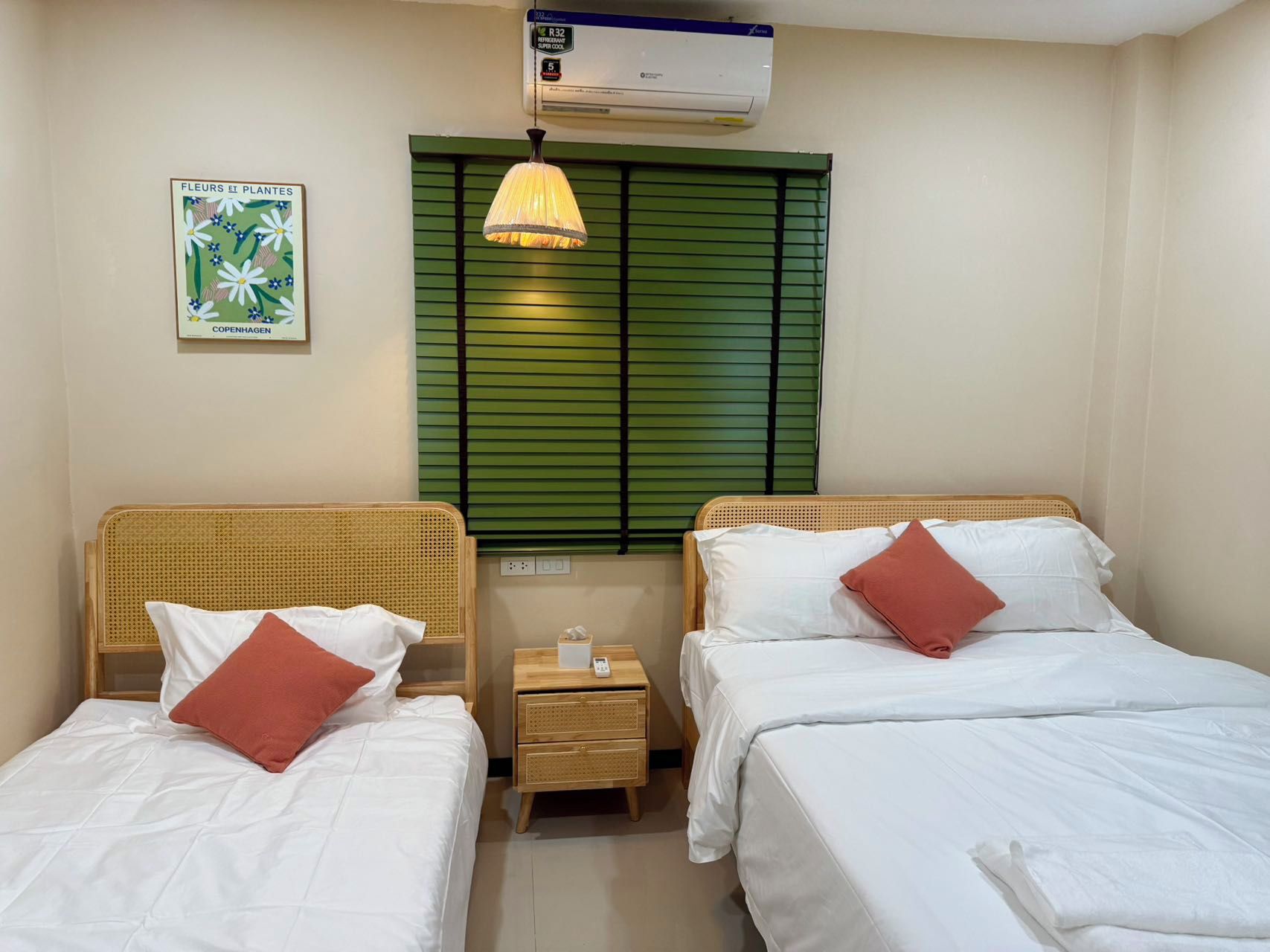 A hotel room with two single beds, a bedside table with a lamp, a wall-mounted air conditioner, and a framed picture.