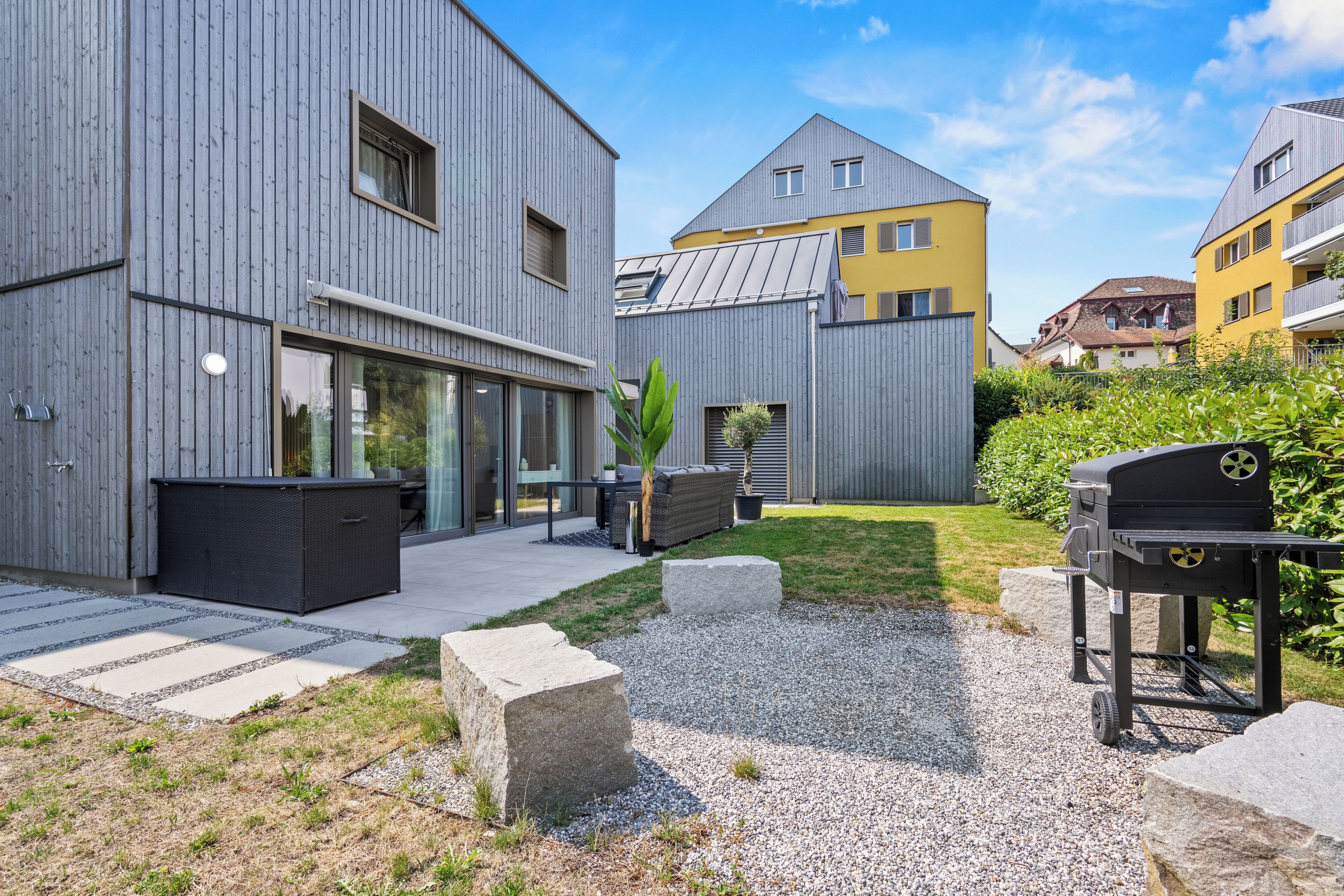 A modern residential building with a patio area, outdoor seating, and a barbecue grill.