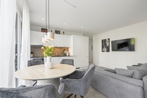 A modern living space with a dining area, grey upholstered sofa, and a flat-screen TV mounted on the wall.