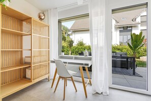 Een moderne kamer met een groot raam, een houten boekenkast, een wit bureau met stoel en uitzicht op een woning en tuin.