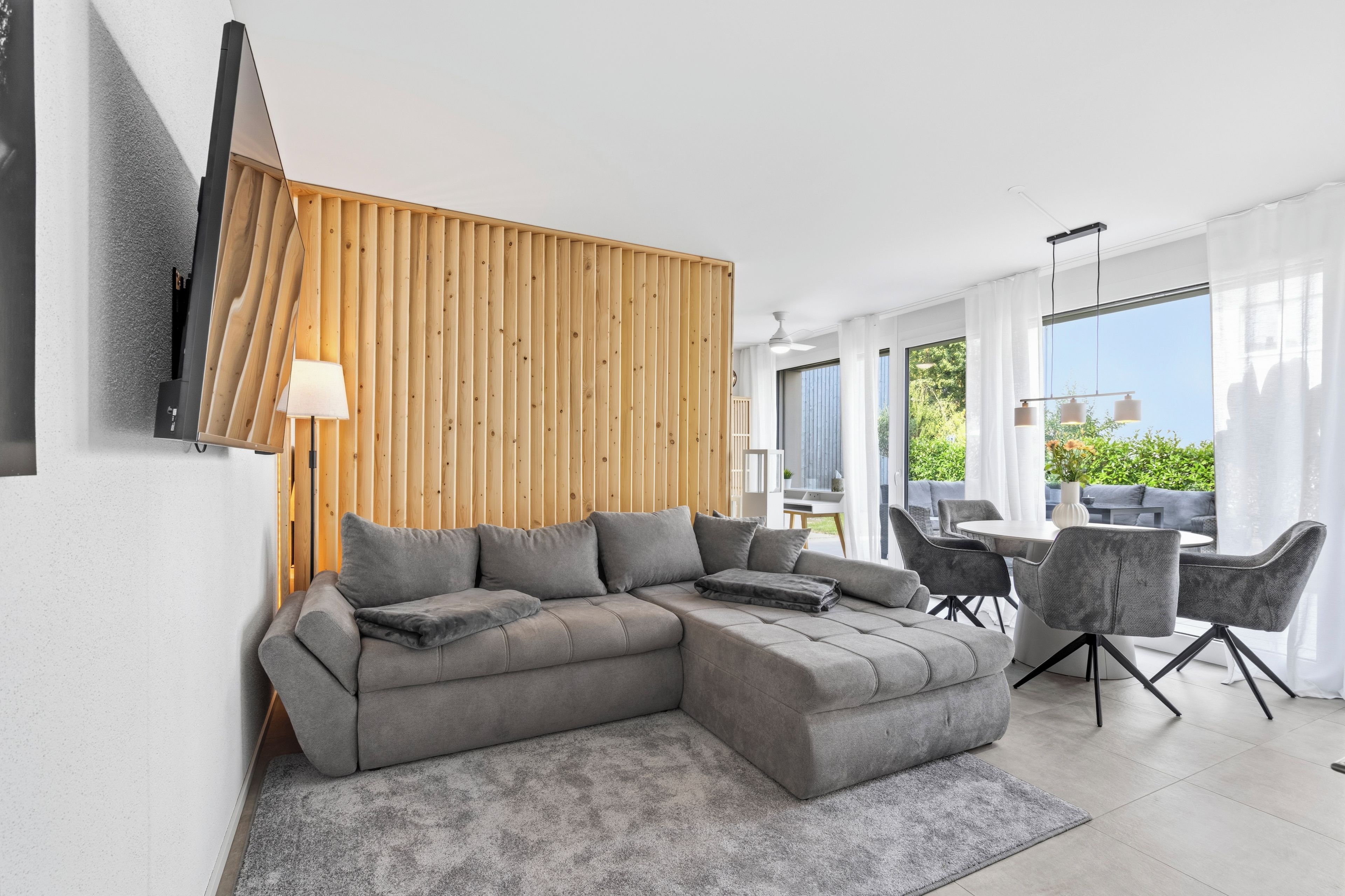 Un salon moderne avec un canapé d’angle, un mur d’accent en bois, un téléviseur à écran plat fixé au mur et un coin repas avec une table et des chaises.