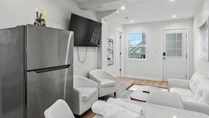 A modern living room with a large stainless steel refrigerator, white sofas, a flat-screen TV, and a window with a view of a building. - On the Pier Cottage with Direct Beach Access (Santa Monica)
