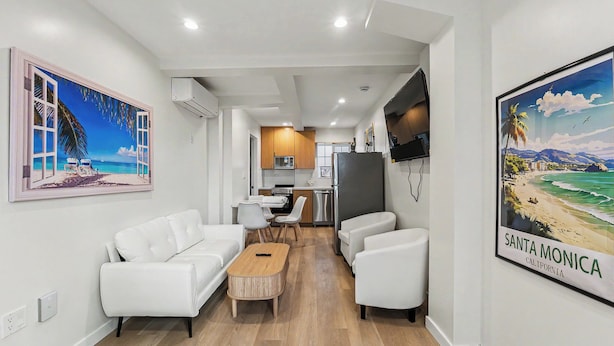 A modern living room with a sofa, chairs, a coffee table, and a TV. There is a kitchen area with cabinets and appliances. - On the Pier Cottage with Direct Beach Access (Santa Monica)
