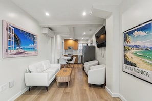 A modern living room with a sofa, chairs, a coffee table, and a TV. There is a kitchen area with cabinets and appliances.