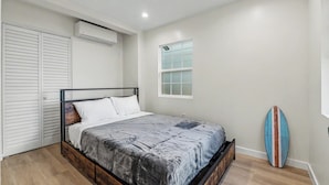 A bedroom with a bed, a wardrobe, an air conditioning unit, a window, and a surfboard. - On the Pier Cottage with Direct Beach Access (Santa Monica)