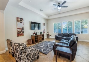 A spacious living room with a ceiling fan, a large flat-screen TV, a sectional sofa, a loveseat, and a coffee table.