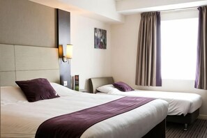A hotel room with two beds, a headboard, a bedside table, a lamp, and a window with curtains.