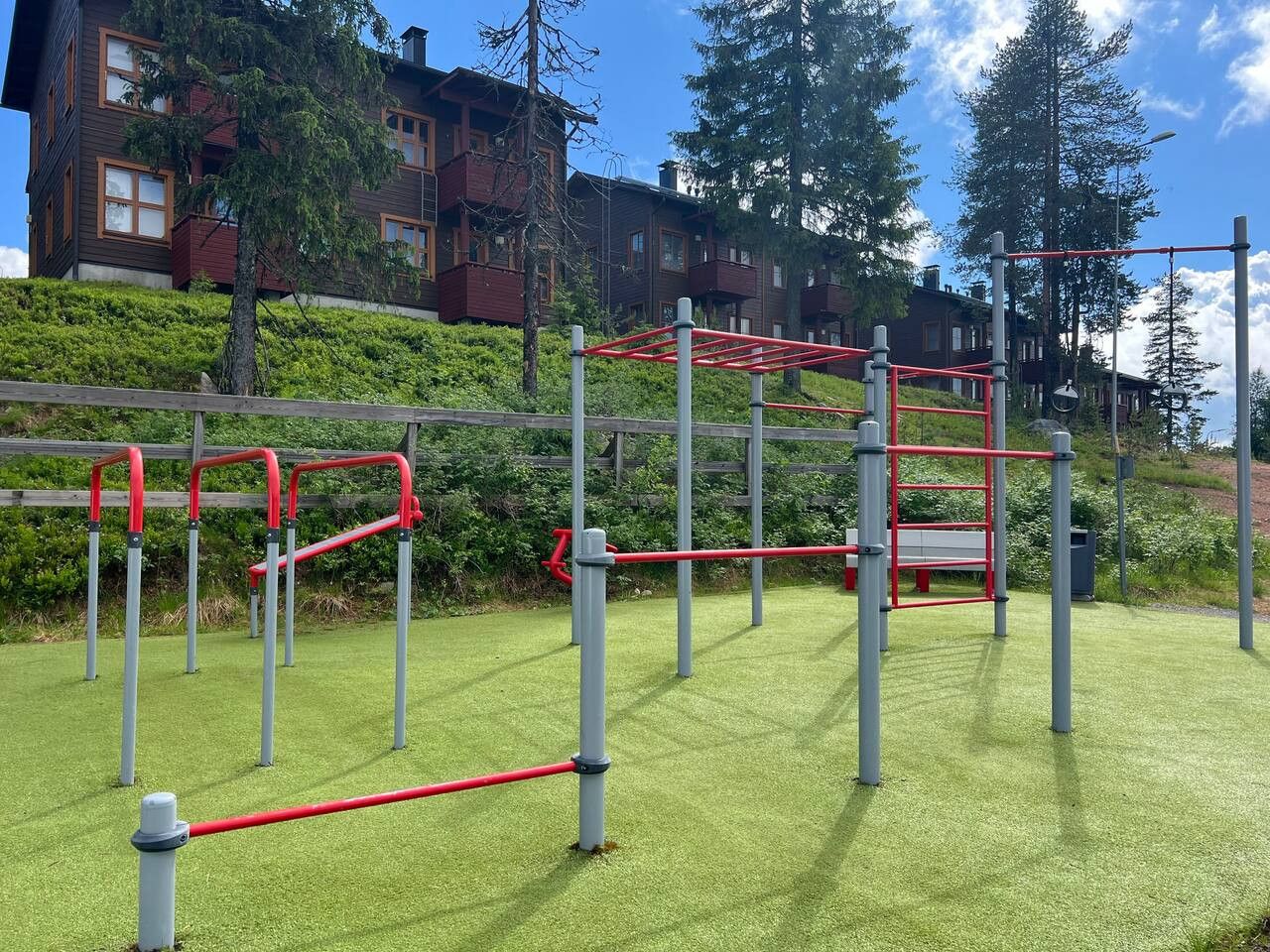 Un gimnasio al aire libre con equipo rojo y gris sobre césped artificial verde, rodeado de árboles y edificios residenciales.