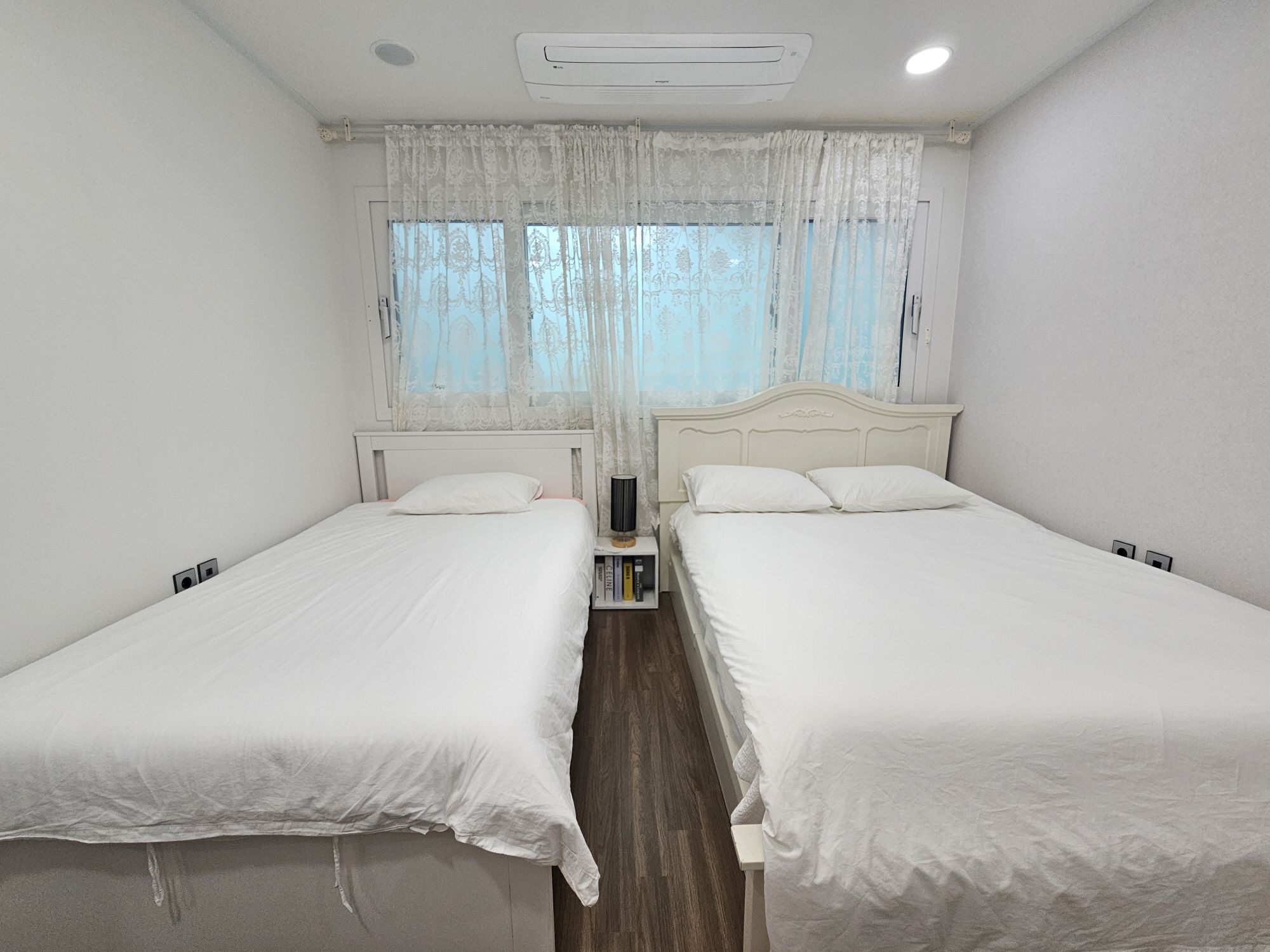 A room with two single beds, a window with sheer curtains, a bedside table with books, and a ceiling air conditioner.