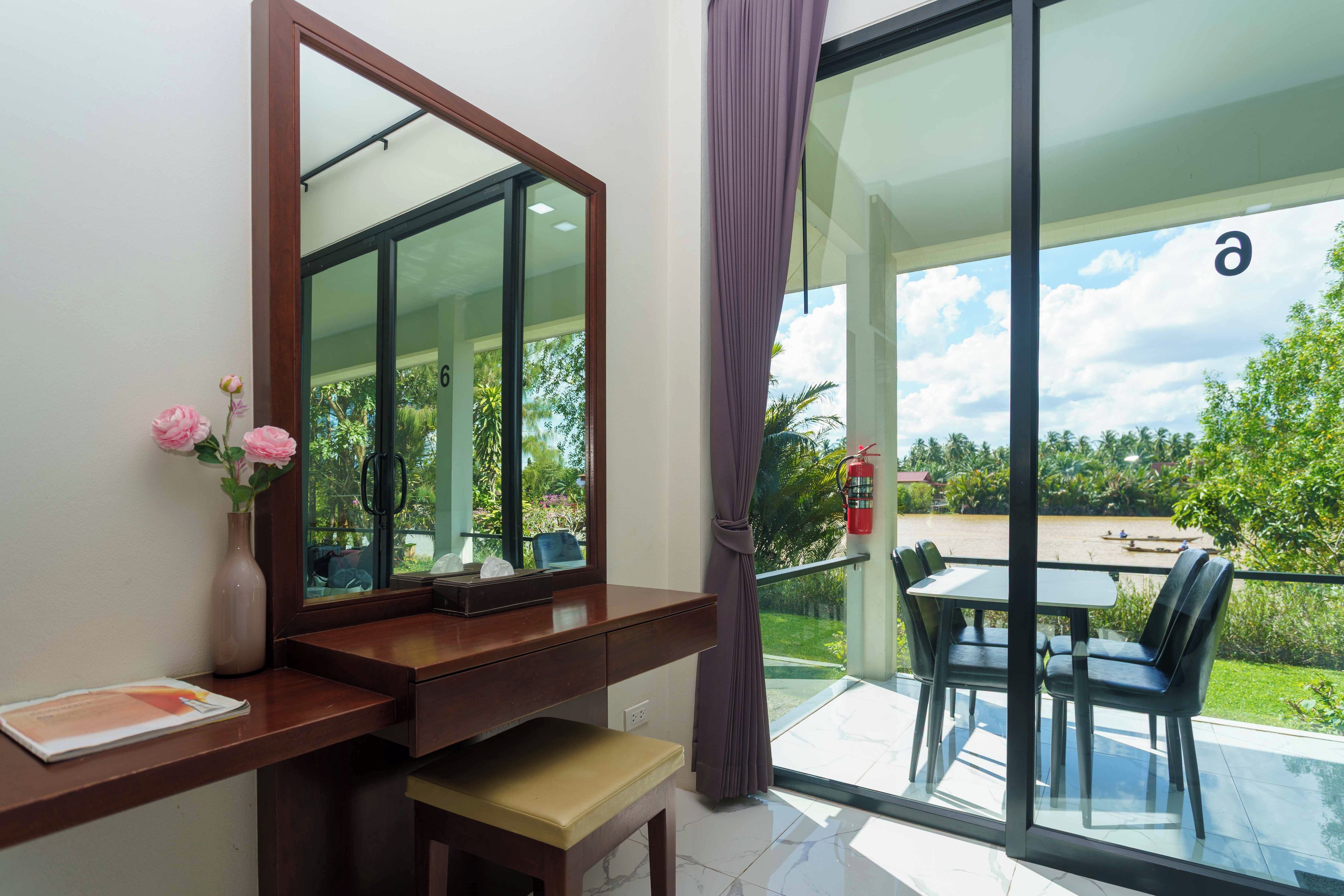 A room with a large mirror, a desk with a chair, a small bench, and a view of a balcony with a table and chairs.