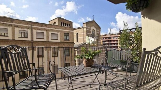 Een balkon met metalen stoelen en een tafel, een bloempot en gebouwen op de achtergrond.