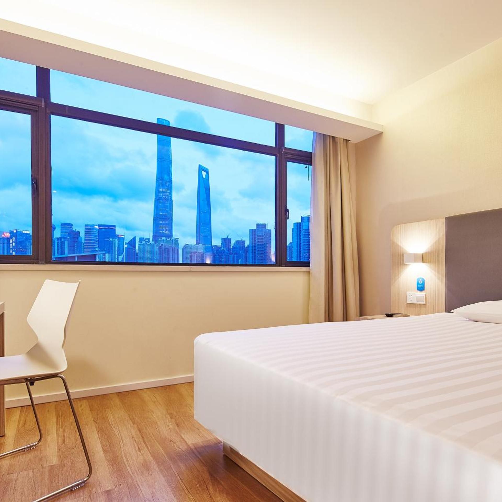 A hotel room with a large window, a bed, a chair, and a wall-mounted light.