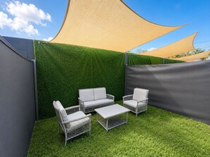 Ein Außenbereich mit Sitzgelegenheit, einem Tisch und vier Stühlen, umgeben von einer grünen Hecke und einem großen Sonnensegel.