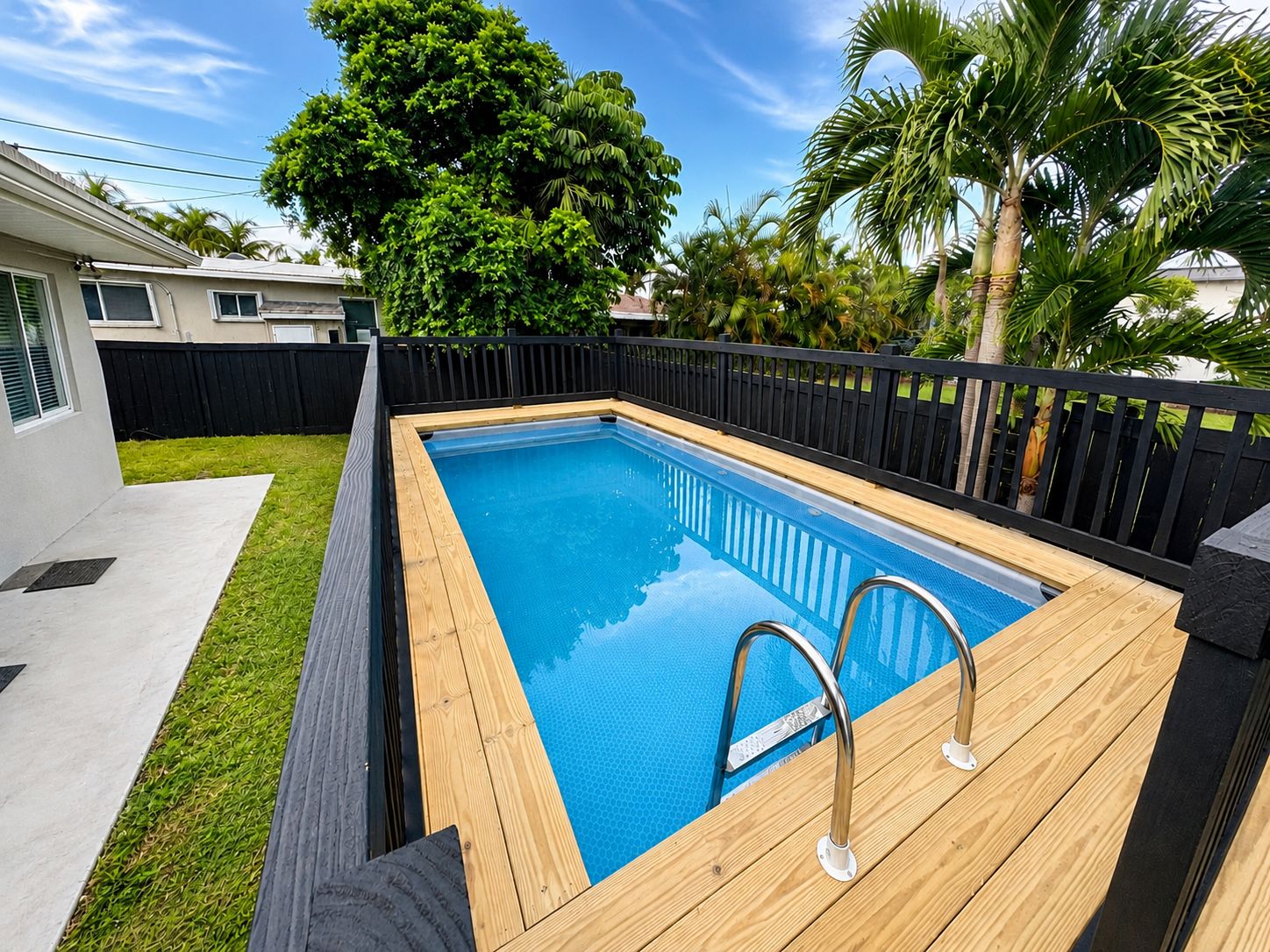 Une piscine rectangulaire dotée d’une terrasse en bois et d’un escalier en métal, entourée d’une clôture noire et d’une végétation luxuriante.