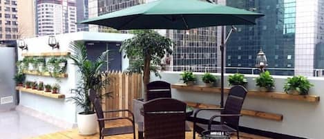 Un área de estar al aire libre con una mesa, sillas, un paraguas verde y plantas en macetas.