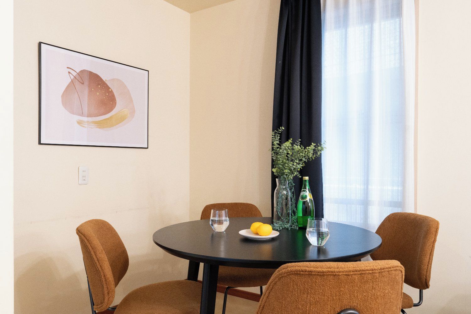 A dining area with a round table, four chairs, a framed artwork, a vase with plants, and a bottle.