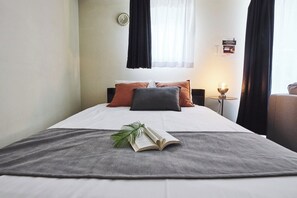 Hotellihuone, jossa on sänky, tyynyjä, yöpöytä lampun kanssa ja ikkuna, jossa on verhot.
