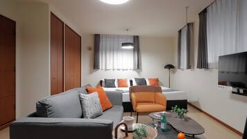 A modern hotel room with a sofa, armchair, dining table, and a bed with a TV.