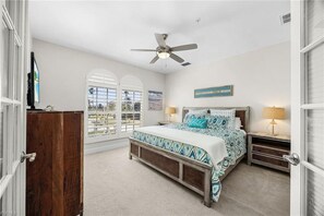 Ein Schlafzimmer mit einem Bett, einem Deckenventilator, einem Nachttisch, einer Lampe und Blick durch das Fenster.