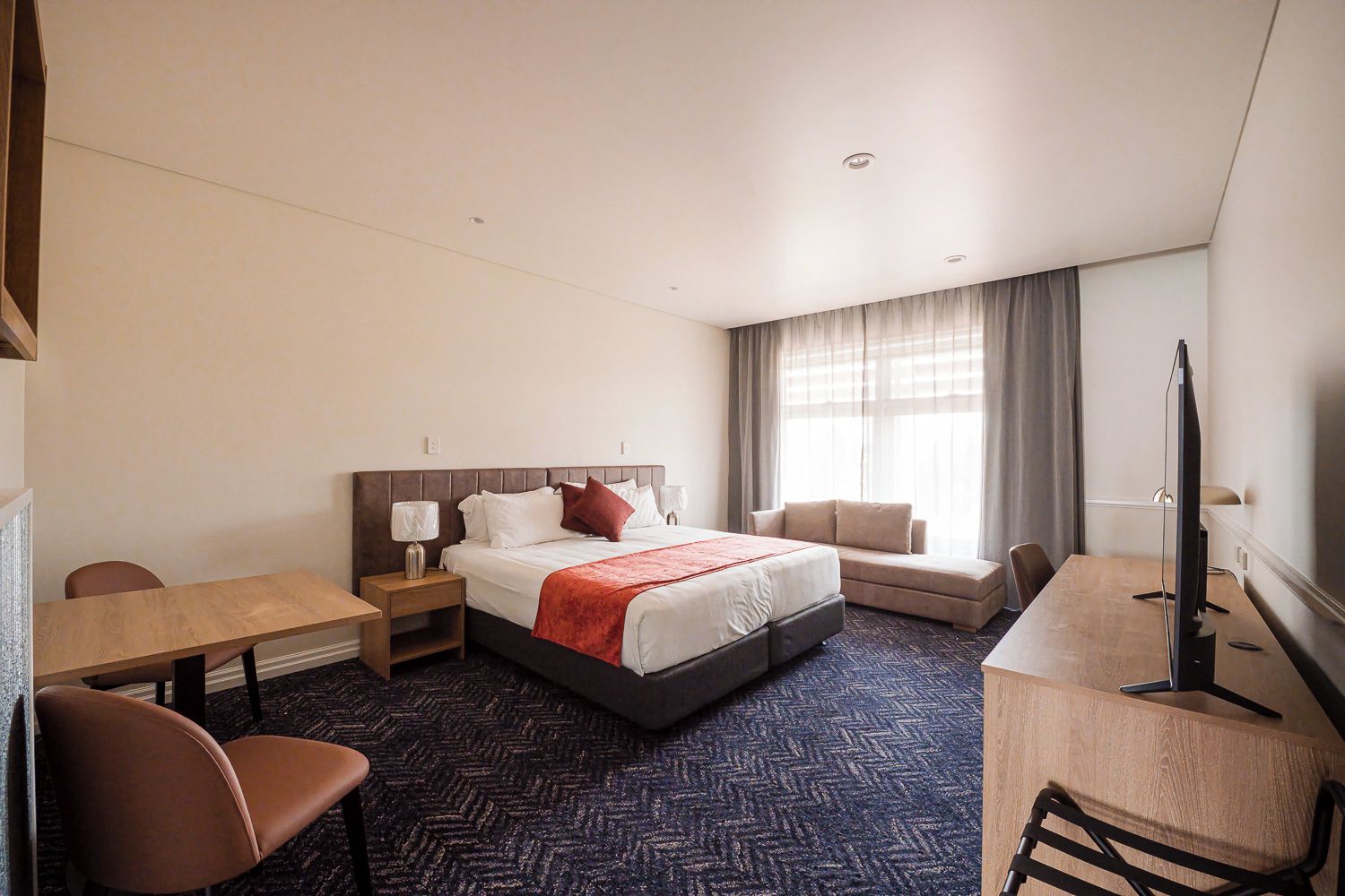 Una habitación de hotel con cama, escritorio, silla, televisor y ventana con cortinas.