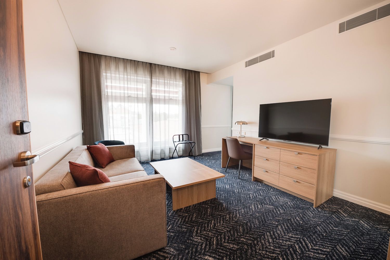 A hotel room with a sofa, a coffee table, a TV on a wooden cabinet, and a desk with a chair.