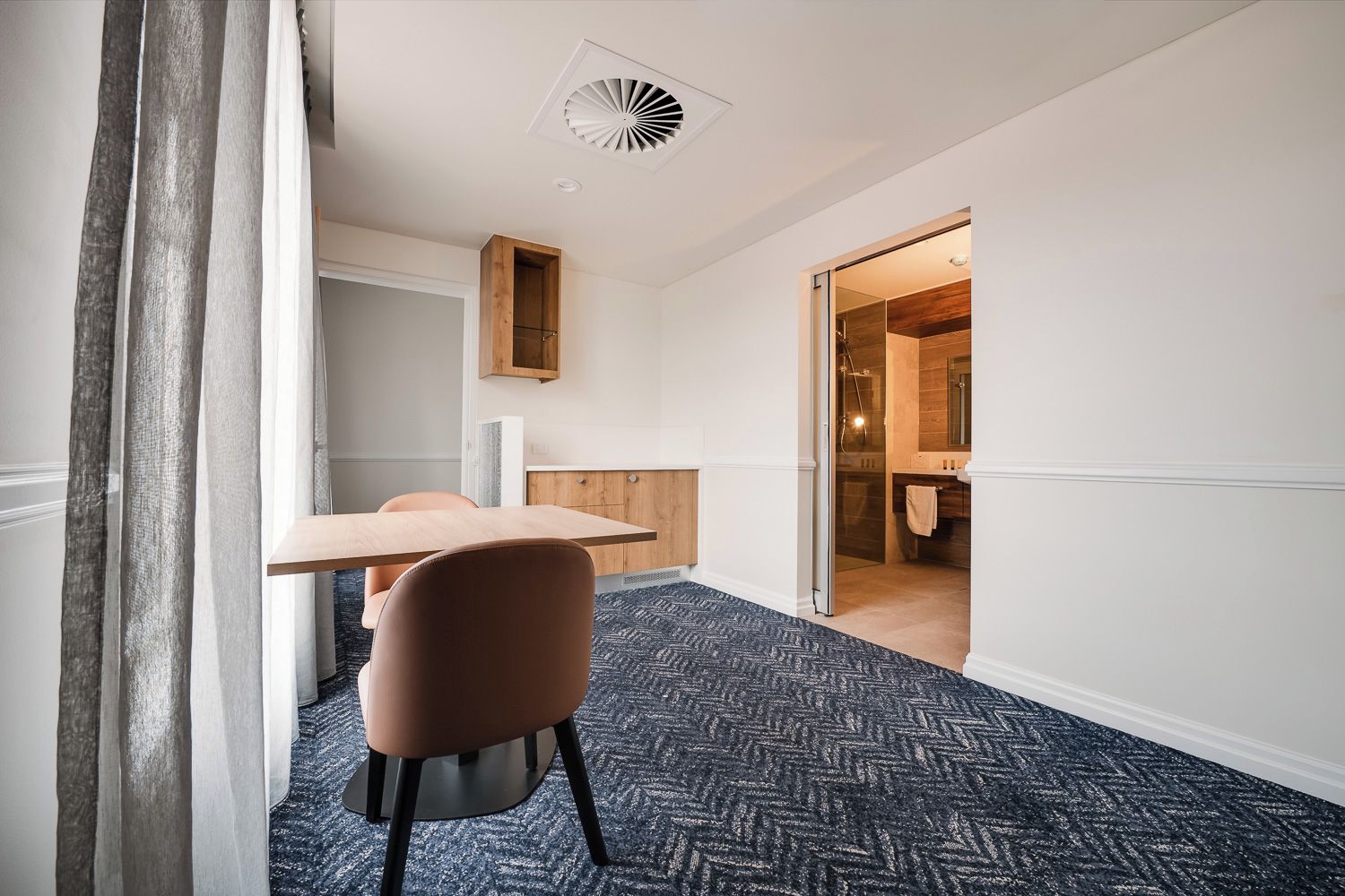 Habitación de hotel con una mesa de madera, una silla, alfombra estampada y vista al baño.
