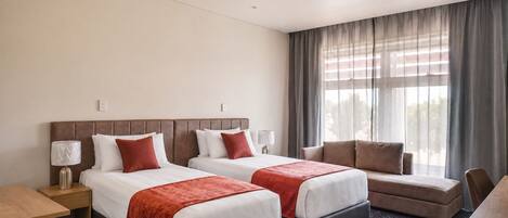 A hotel room with two beds, a sofa, a desk, and a large window with curtains.
