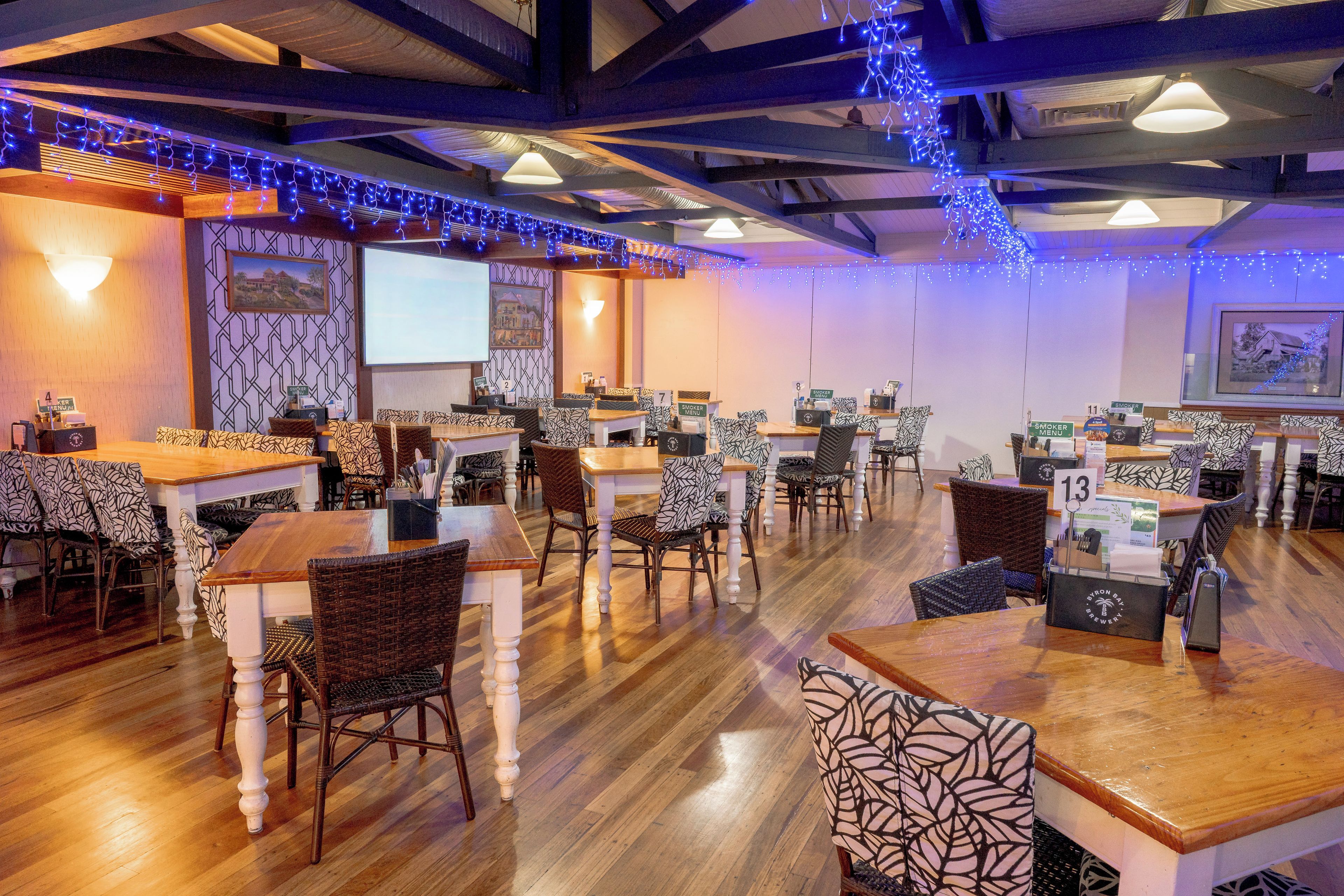 A restaurant interior with wooden tables and chairs, a projector screen, and blue string lights.