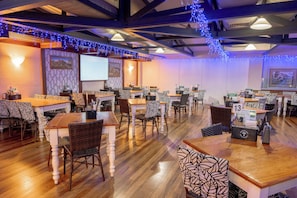 Interior de un restaurante con mesas y sillas de madera, una pantalla de proyector y luces azules tipo guirnalda.