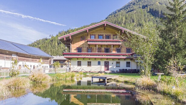 Edifício de madeira de dois andares refletido em um lago, cercado por vegetação e montanhas.
