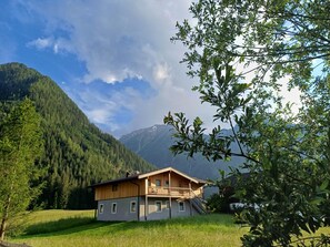 A wooden cabin with a balcony, surrounded by a forest and mountains.