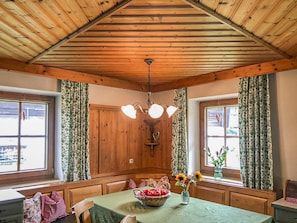Una habitación con vigas de madera en el techo, una araña, una mesa con mantel verde y cortinas florales.