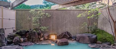 A hot spring area with a wooden pergola overhead, surrounded by rocks and a concrete wall.