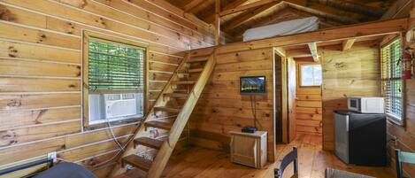 A cabin interior with a wooden staircase, a television mounted on the wall, and a small kitchenette area.
