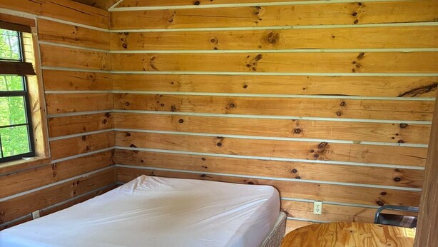 Interior de uma cabana de madeira com uma cama, uma mesa e uma cadeira.