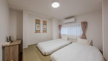 A hotel room with two single beds, a wooden desk, a small trash bin, and a wall-mounted air conditioner.