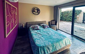 Un dormitorio con una cama grande, una mesita de noche, una silla de mimbre y vistas al exterior a través de una puerta corredera.