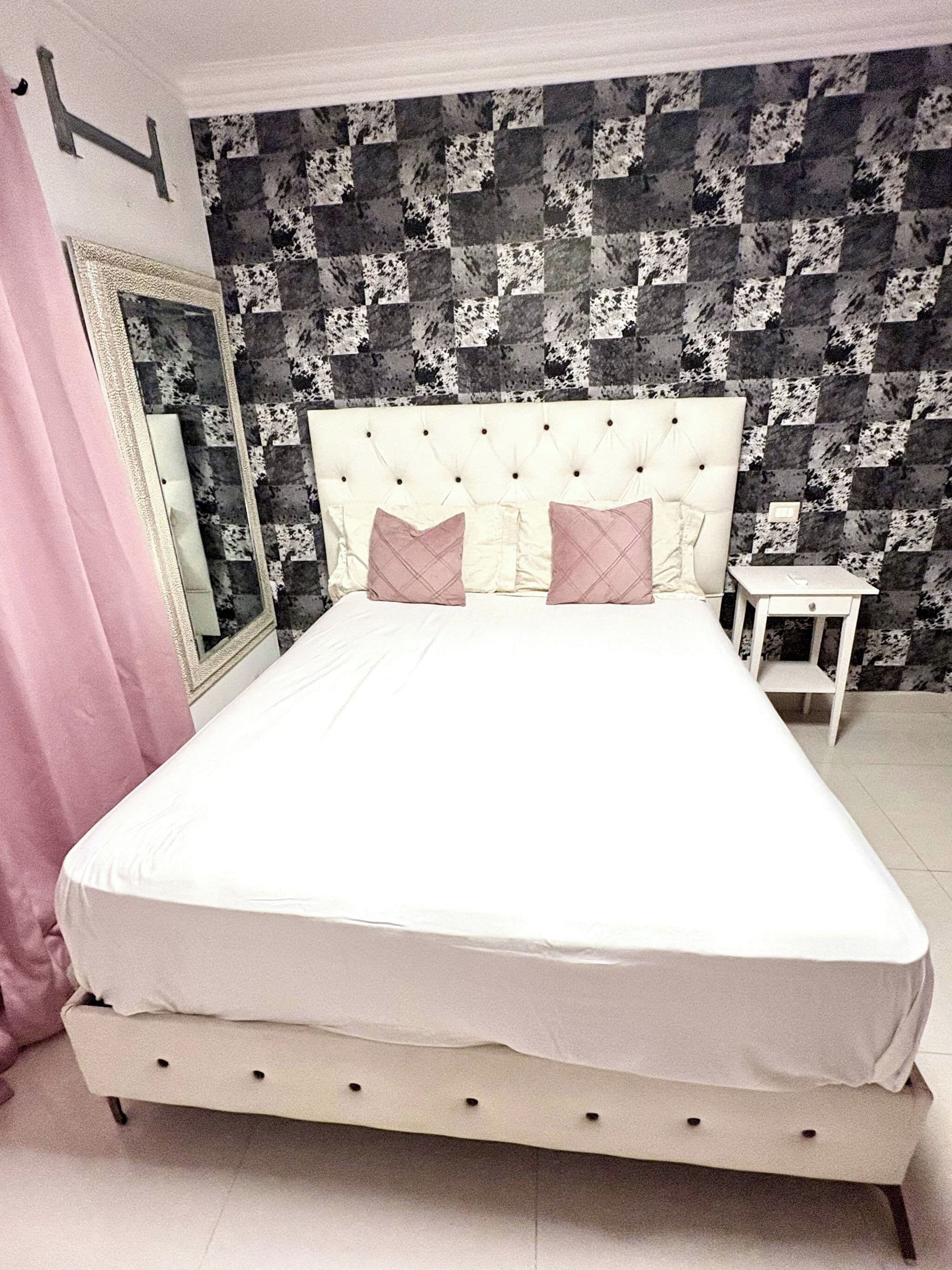 A bed with a white headboard and two pillows, a bedside table, and a mirror on the wall.