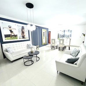 A modern living room with a white sofa, a round coffee table, and a dining area with chairs and a table. - Spacious 3BR Near Airport & Ocean (Santo Domingo Este)