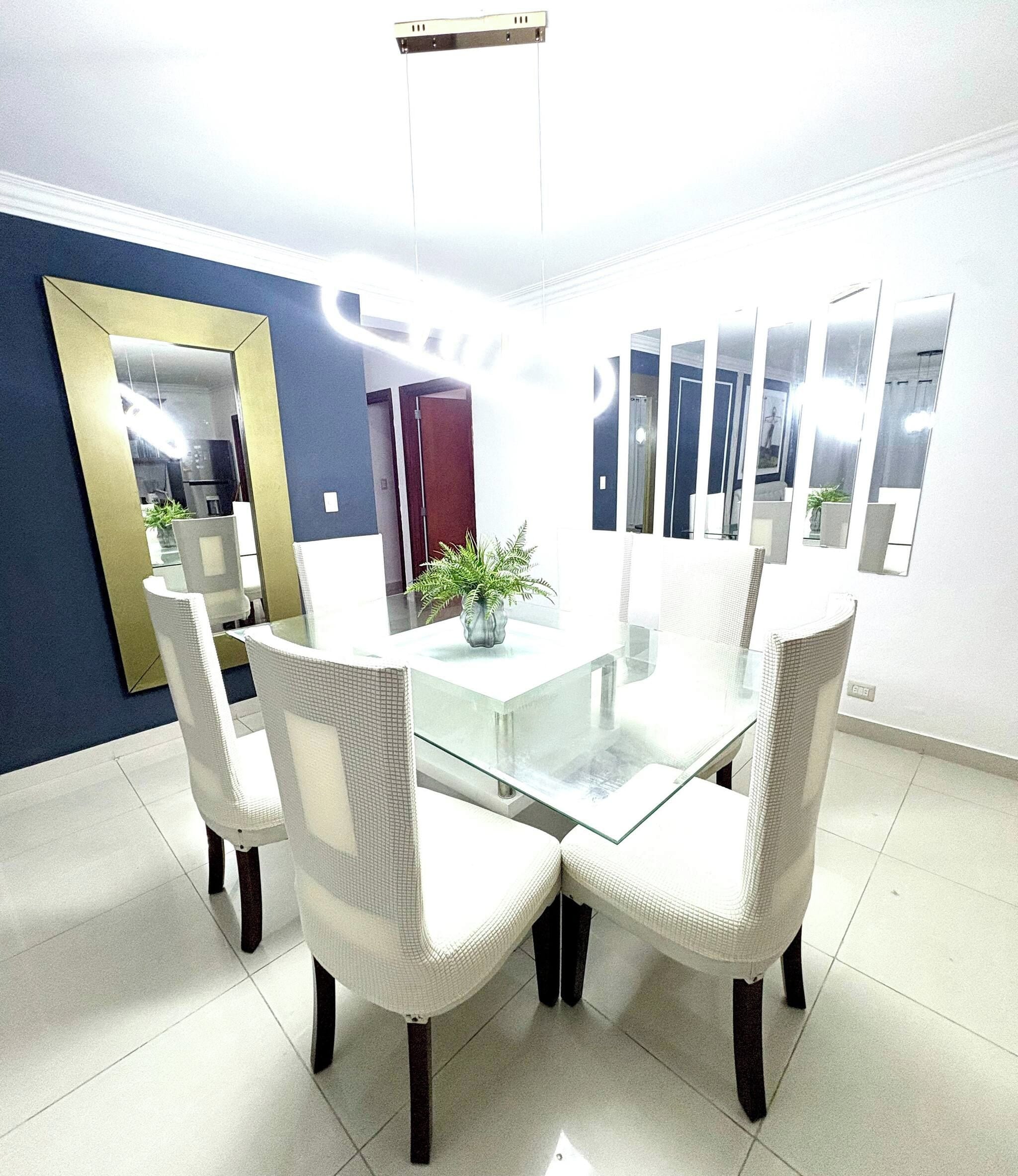 A dining area with a glass-top table surrounded by white chairs, a potted plant, and a large mirror on the wall.