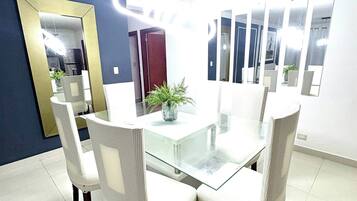 A dining area with a glass-top table surrounded by white chairs, a potted plant, and a large mirror on the wall.