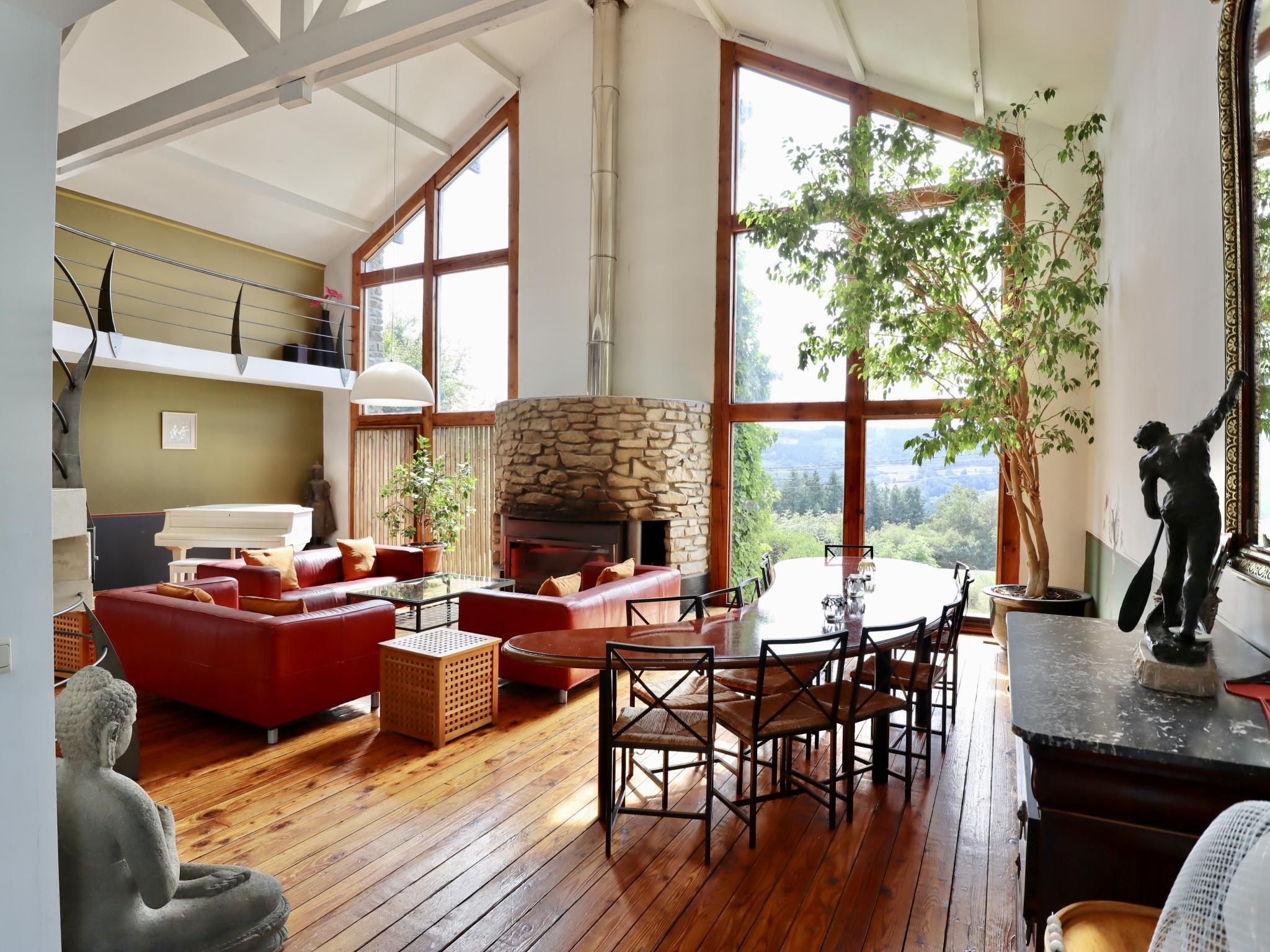 A spacious living room with a stone fireplace, red sofas, wooden flooring, and large windows offering a view of the outdoors.