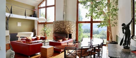A spacious living room with a stone fireplace, red sofas, wooden flooring, and large windows offering a view of the outdoors.