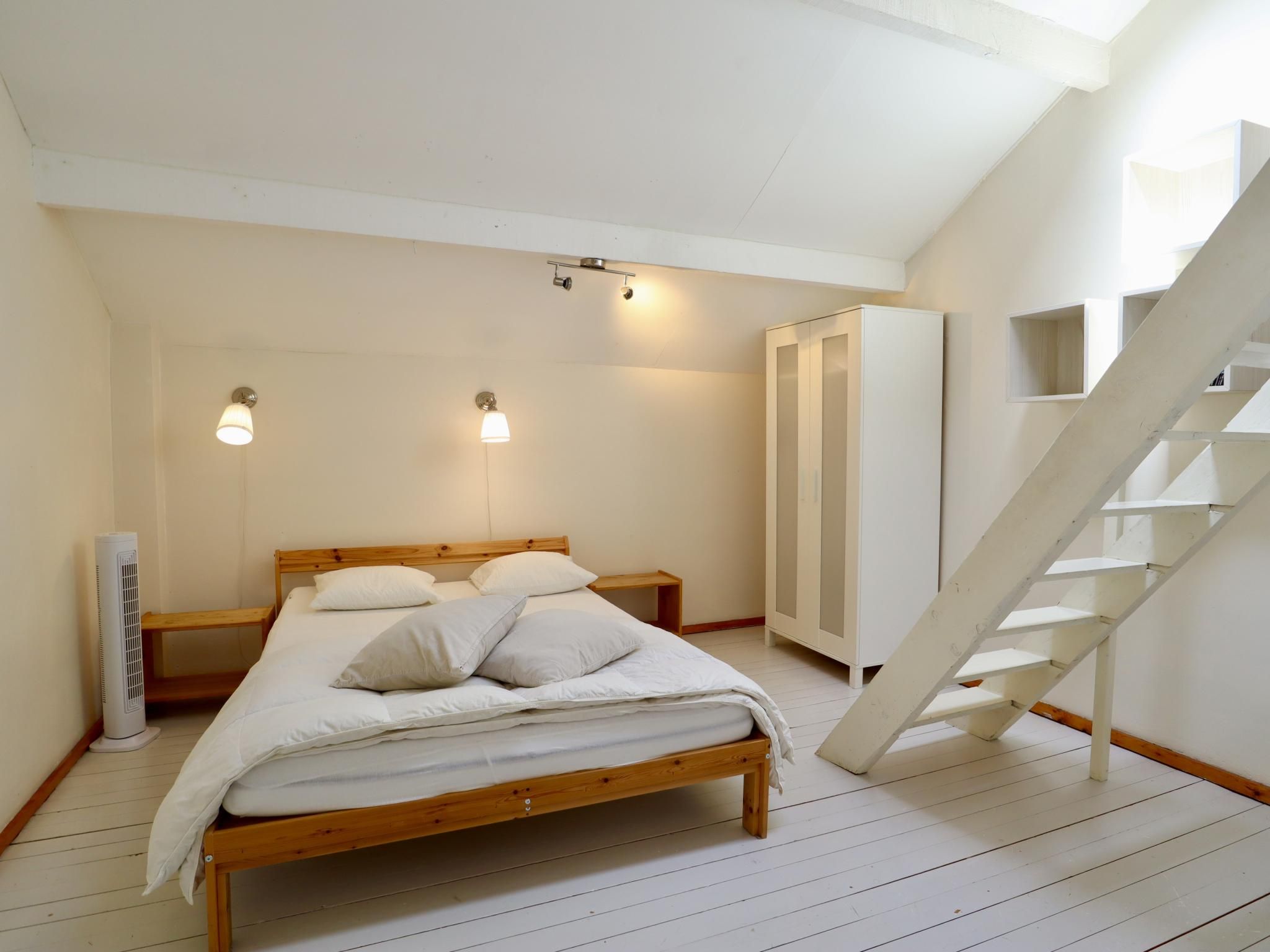 Une chambre avec un lit en bois, des murs blancs, une armoire blanche et un escalier blanc.