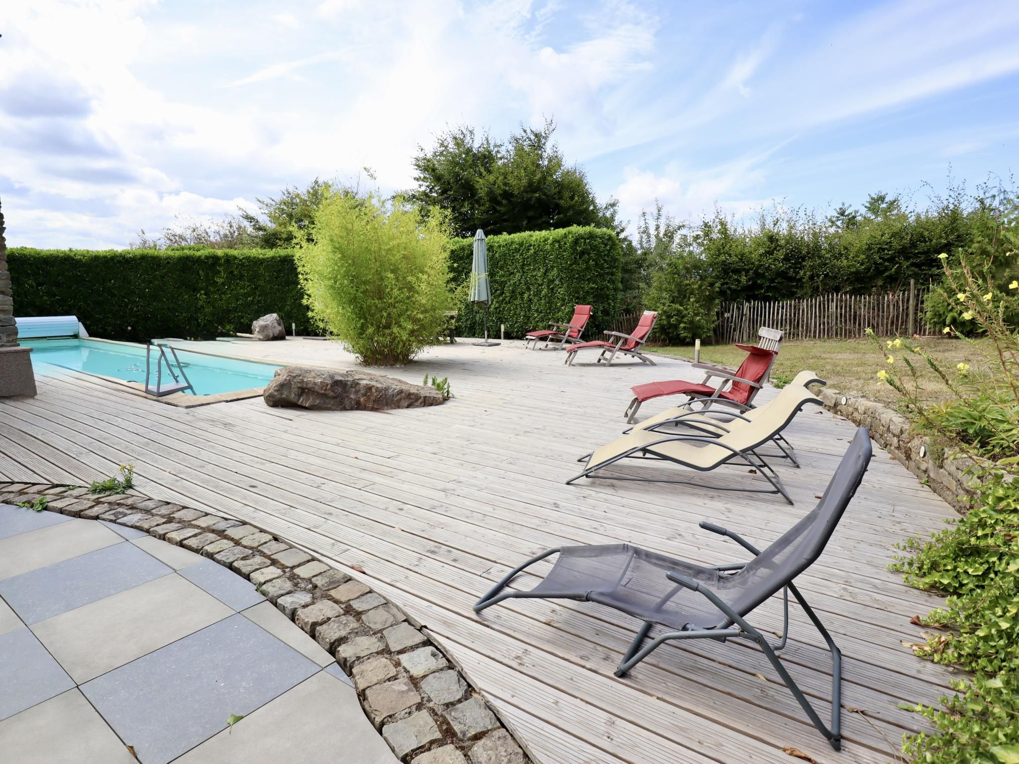 Een houten terras met loungebedden, een zwembad en een haag.