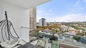 A balcony with a hanging chair, a view of a cityscape with buildings and trees, and a clear sky. - Luxury Sydney Residence with Pool, Gym & City Views (Darlinghurst)