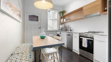 A modern kitchen with white cabinetry, a dining table with chairs, a hanging light fixture, and a window with blinds.